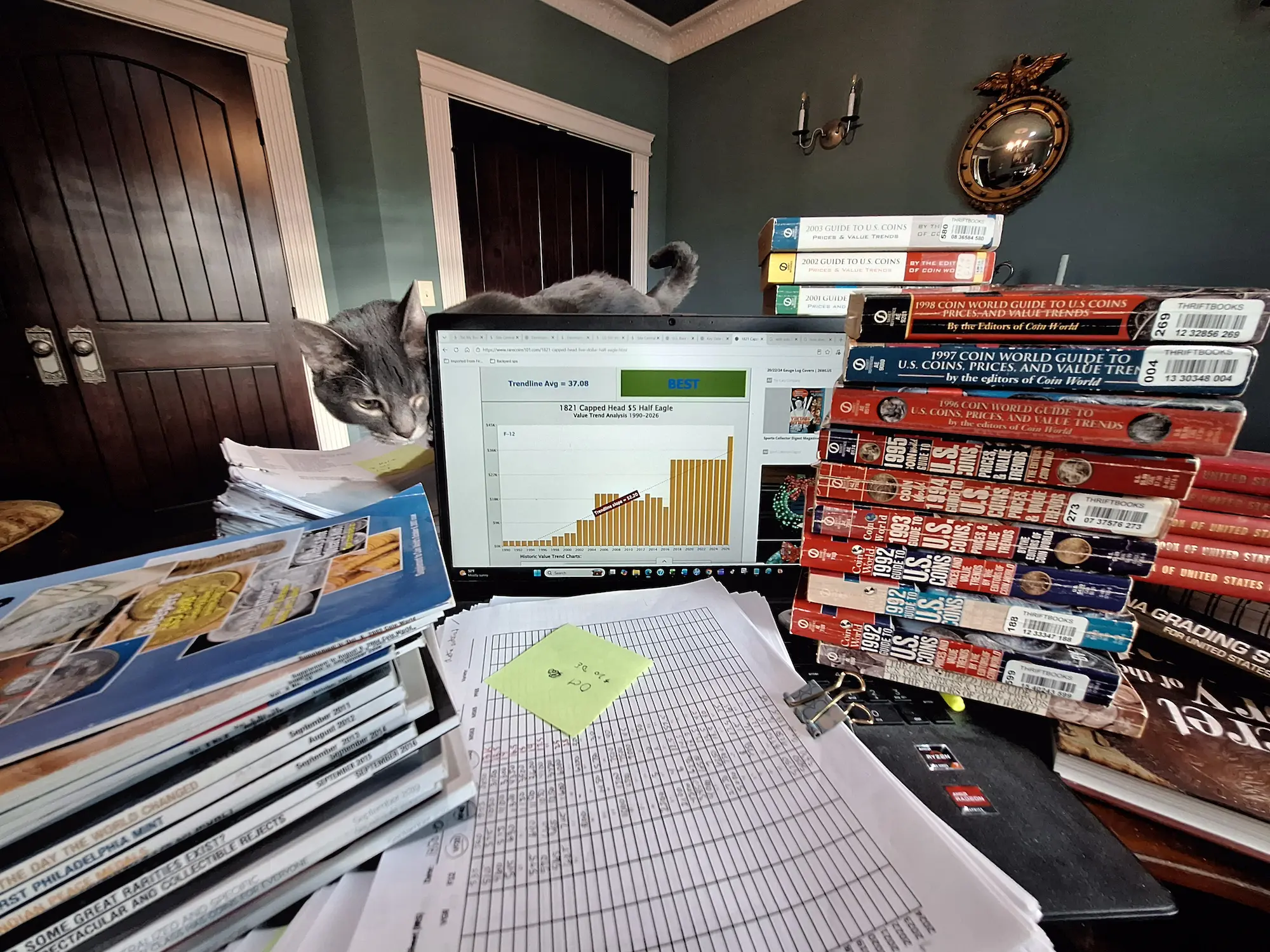 Rare coin value research behind Rare Coins 101 Old coin price guides, handwritten data sheets, a laptop displaying a rare coin value chart, and a cat beside Dan’s research materials for Rare Coins 101.